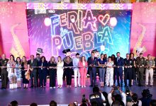 Una toma amplia y nítida de una ceremonia de corte de listón en un gran escenario para la inauguración de la Feria de Puebla 2026. Alrededor de veinte dignatarios, incluidos funcionarios gubernamentales y militares, están alineados sosteniendo un listón