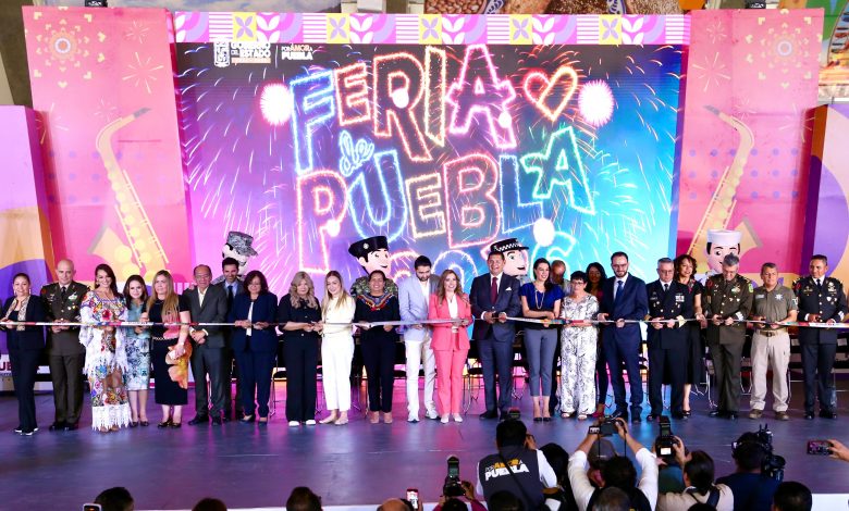 Una toma amplia y nítida de una ceremonia de corte de listón en un gran escenario para la inauguración de la Feria de Puebla 2026. Alrededor de veinte dignatarios, incluidos funcionarios gubernamentales y militares, están alineados sosteniendo un listón