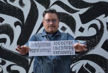 Jesús Barrientos Mora, académico de la BUAP, muestra un cuadernillo con alfabetos de caligrafía gótica frente a un mural de letras góticas en blanco y negro.
