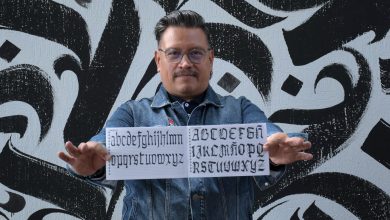 Jesús Barrientos Mora, académico de la BUAP, muestra un cuadernillo con alfabetos de caligrafía gótica frente a un mural de letras góticas en blanco y negro.