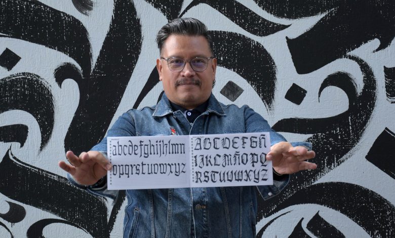 Jesús Barrientos Mora, académico de la BUAP, muestra un cuadernillo con alfabetos de caligrafía gótica frente a un mural de letras góticas en blanco y negro.