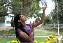 Perla Juárez Filomeno, estudiante de danza de la BUAP, baila al aire libre con los brazos extendidos hacia las ramas de un árbol, vestida con una blusa morada de estampado ondulado.