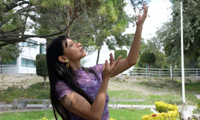 Perla Juárez Filomeno, estudiante de danza de la BUAP, baila al aire libre con los brazos extendidos hacia las ramas de un árbol, vestida con una blusa morada de estampado ondulado.